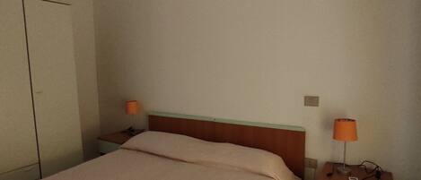 A simple, clean hotel room with a bed, a nightstand with a lamp, a wardrobe, and an air conditioning unit on the wall.