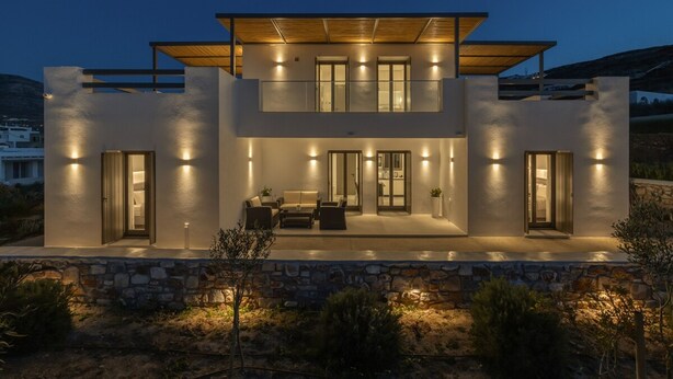 A modern two-story house with a stone foundation, a covered patio, and a well-lit interior. - Cozy Haven -island Living Paros (Paros)