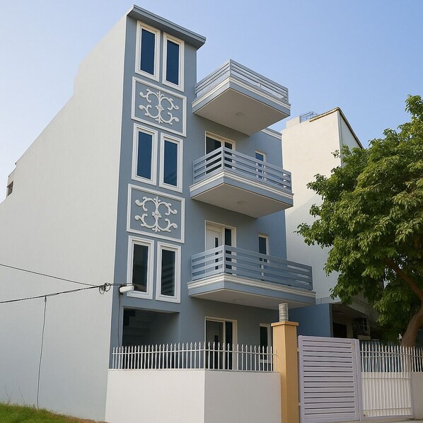 A multi-story residential building with balconies, a white picket fence, and a tree in the background. - The Gleam Hotel (Gurugram)