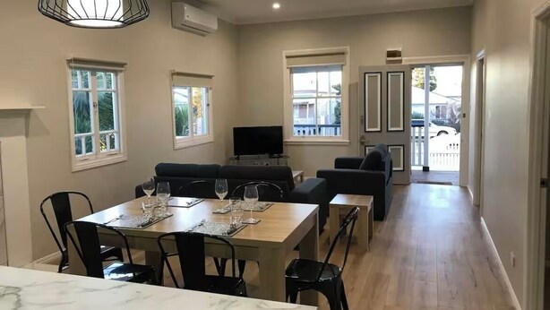 A spacious living area with a marble dining table, black chairs, and a television. There are windows with blinds, a hallway, and a door leading outside. - Eton Cottage: Central Boutique Stay (East Toowoomba)