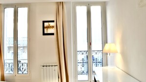 Una habitació d'hotel amb un llit, un radiador, una finestra amb cortina, un quadre a la paret i una làmpada sobre una taula de nit.