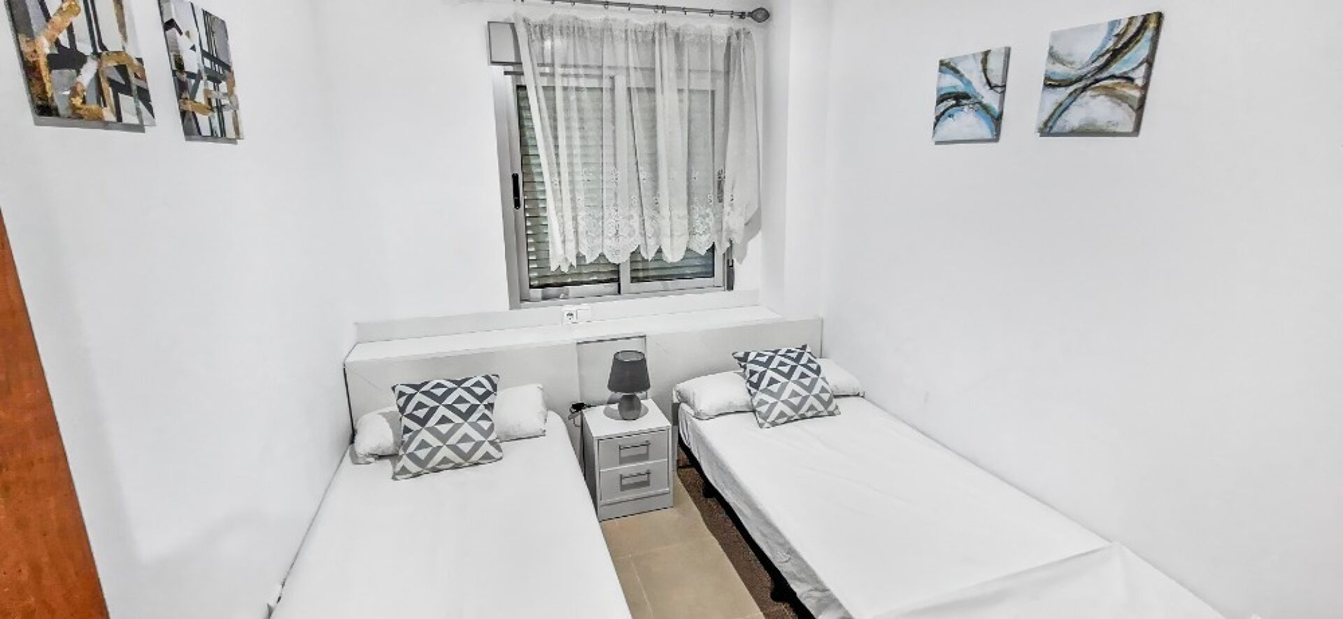 A small, clean room with two single beds, a window with curtains, and a bedside table with a lamp.