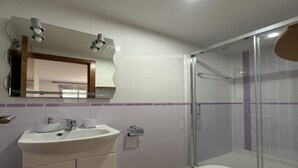 Une salle de bain moderne avec une douche à l’italienne en verre, un lavabo blanc avec un miroir au-dessus et un meuble beige doté de tiroirs.