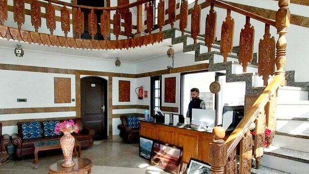 Un vestíbulo de hotel con una recepción, una escalera con barandillas decoradas, un conjunto de sofás y un jarrón con flores.