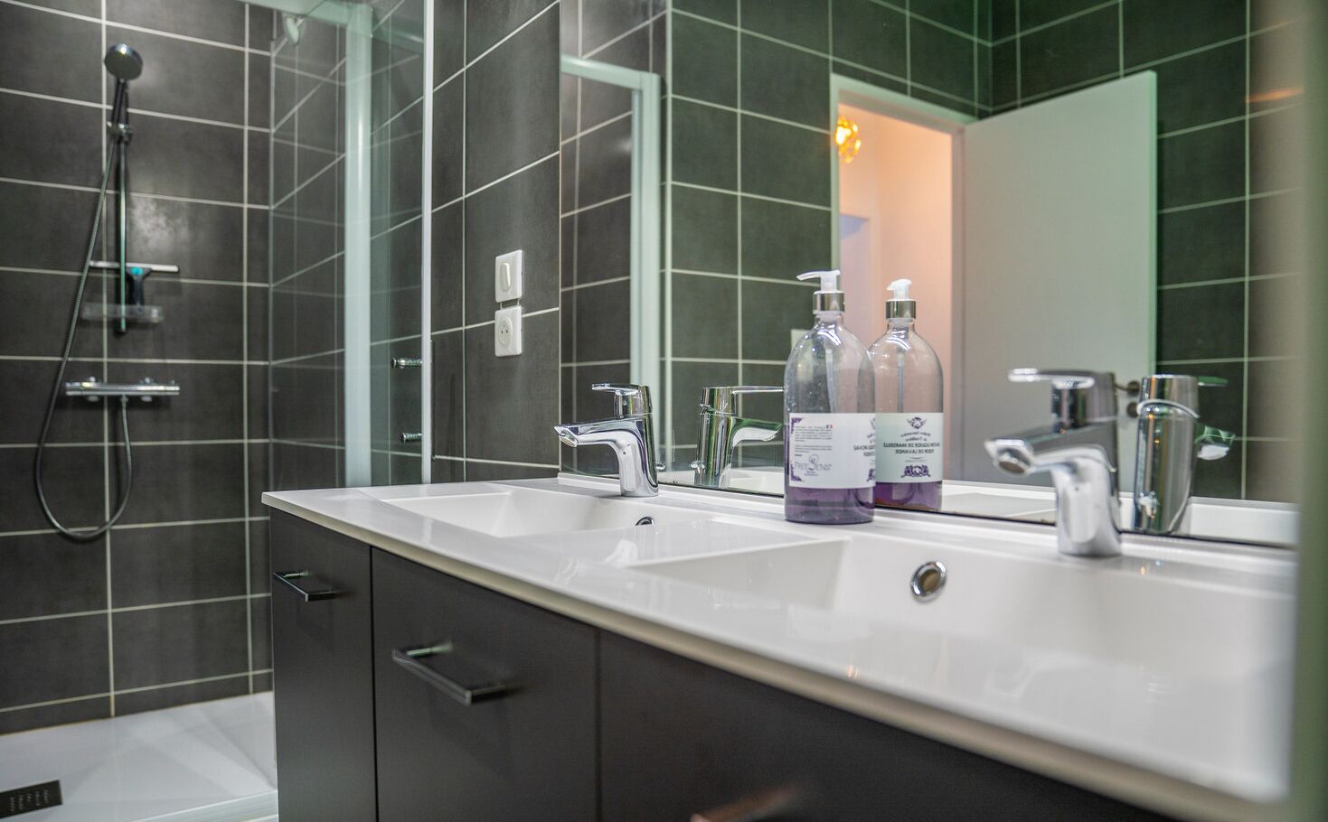 A modern bathroom with a dual sink vanity, a handheld showerhead, and two bottles of hand soap.