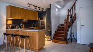 A modern kitchen with wooden cabinets, a central island with stools, a built-in oven, and a staircase with wooden railings.