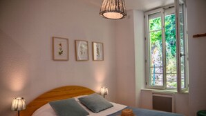 A bedroom with a wooden headboard, a bed with blue bedding, two pillows, a hat on the bed, a window with a view of greenery, and three framed pictures on the wall.