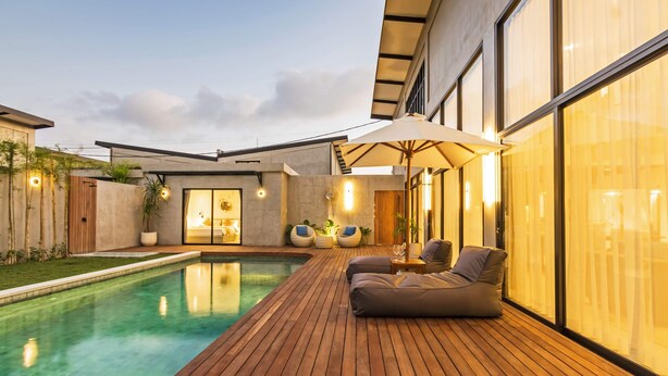 Un área de piscina moderna al aire libre con cubierta de madera, sillas de descanso y un amplio paraguas.