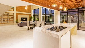 Una cocina moderna con encimera de mármol y estufa integrada. Hay una zona de comedor con una mesa y sillas, y una sala de estar con sofá y televisor.