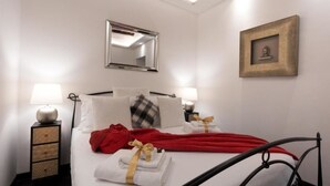 A neatly made bed with white linens, a red blanket, and a red robe. There are two bedside lamps, a framed picture on the wall, and a mirror above the bed. - Barcaccia Luxury Suites: Suite Queen 3 (Roma)