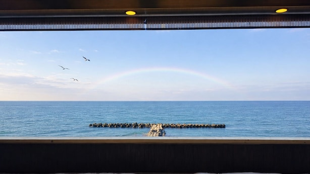 Habitació amb una finestra gran que dona a un mar tranquil, una terrassa de fusta i dues zones d'seients amb coixins.