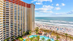 A high-rise hotel with multiple balconies overlooking a beach and a pool area.