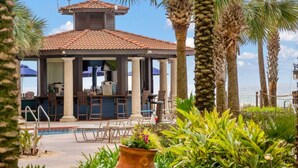 A covered outdoor bar area with a pool, surrounded by palm trees and plants.