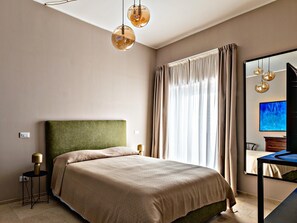 A modern bedroom with a green upholstered headboard, a large bed with a beige bedspread, a side table with a lamp, and a large window with curtains. - Nido di Noto (Noto)