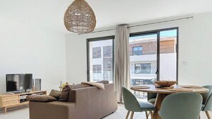 A modern living room with a sofa, a TV, a dining table with chairs, and a large window with a view of buildings.