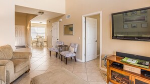 A spacious living room with a sofa, two armchairs, a coffee table, and a TV mounted on the wall. There is a dining area in the background with a table and chairs.