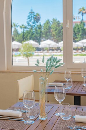 Una mesa dispuesta con copas de vino y un florero con plantas secas, con vistas a sombrillas y árboles al aire libre a través de una ventana.