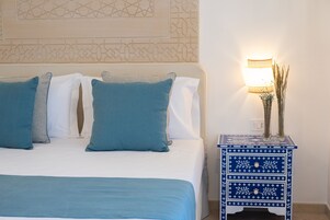Un letto rifatto con lenzuola bianche e azzurre, una lampada da comodino e un vaso decorativo con piante essiccate su un comodino bianco e azzurro.