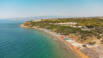 Una playa costera con hileras de sombrillas y tumbonas, un mar azul cristalino y edificios en una colina.