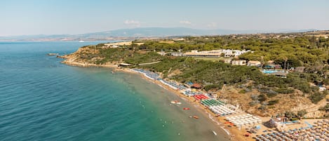 A coastal beach with rows of umbrellas and sun loungers, a clear blue sea, and buildings on a hillside.