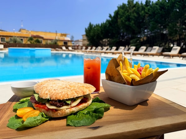A meal with a sandwich, fries, and a drink by a poolside with lounge chairs and a clear sky.
