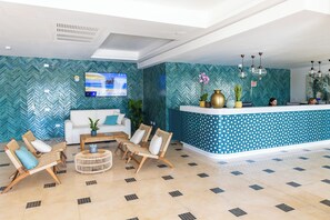 Un área de recepción con un sofá blanco, sillas de mimbre, una mesa de centro y un mostrador de recepción con plantas decorativas.