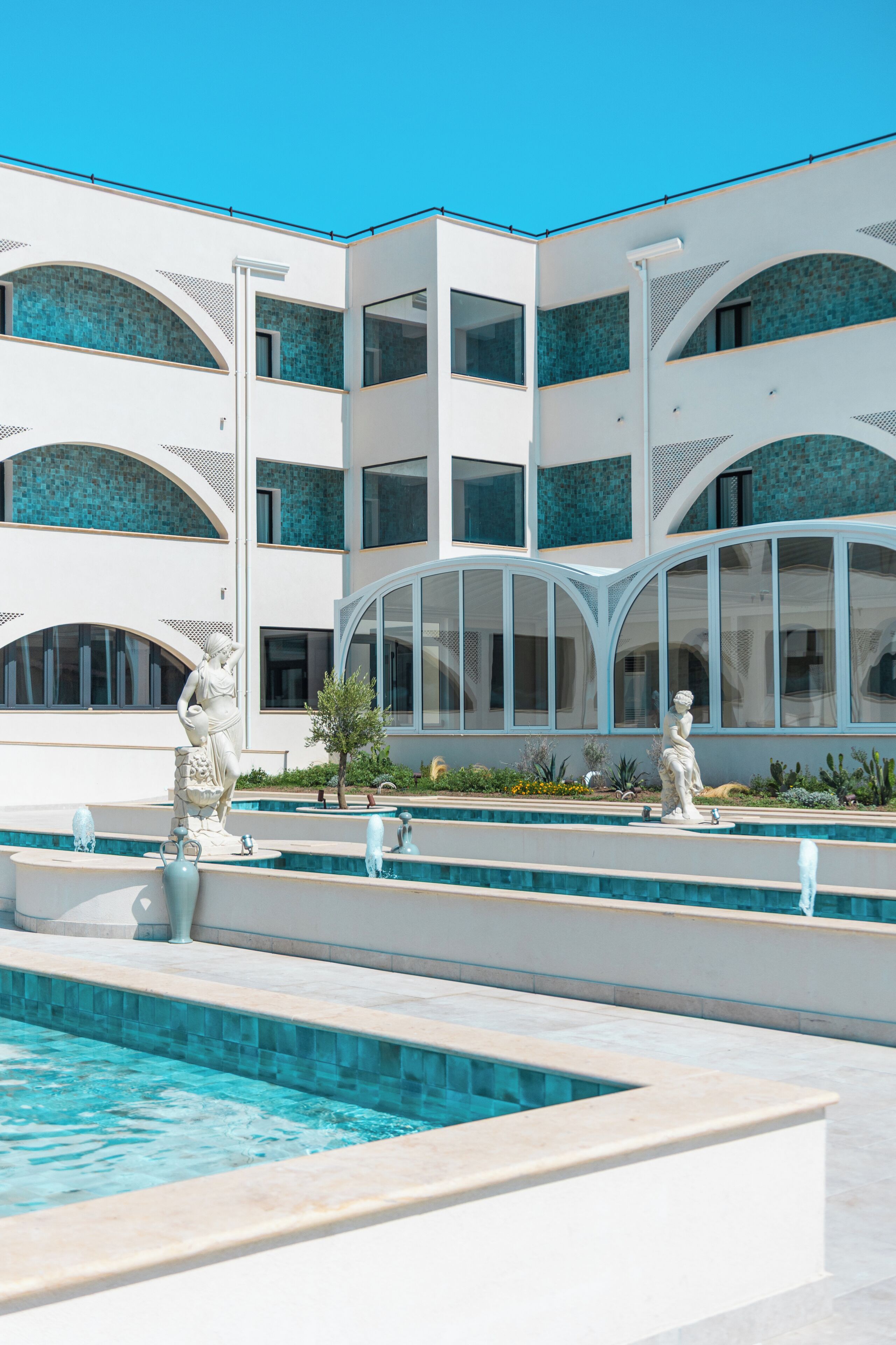 A modern building with multiple balconies, a swimming pool, and classical statues.