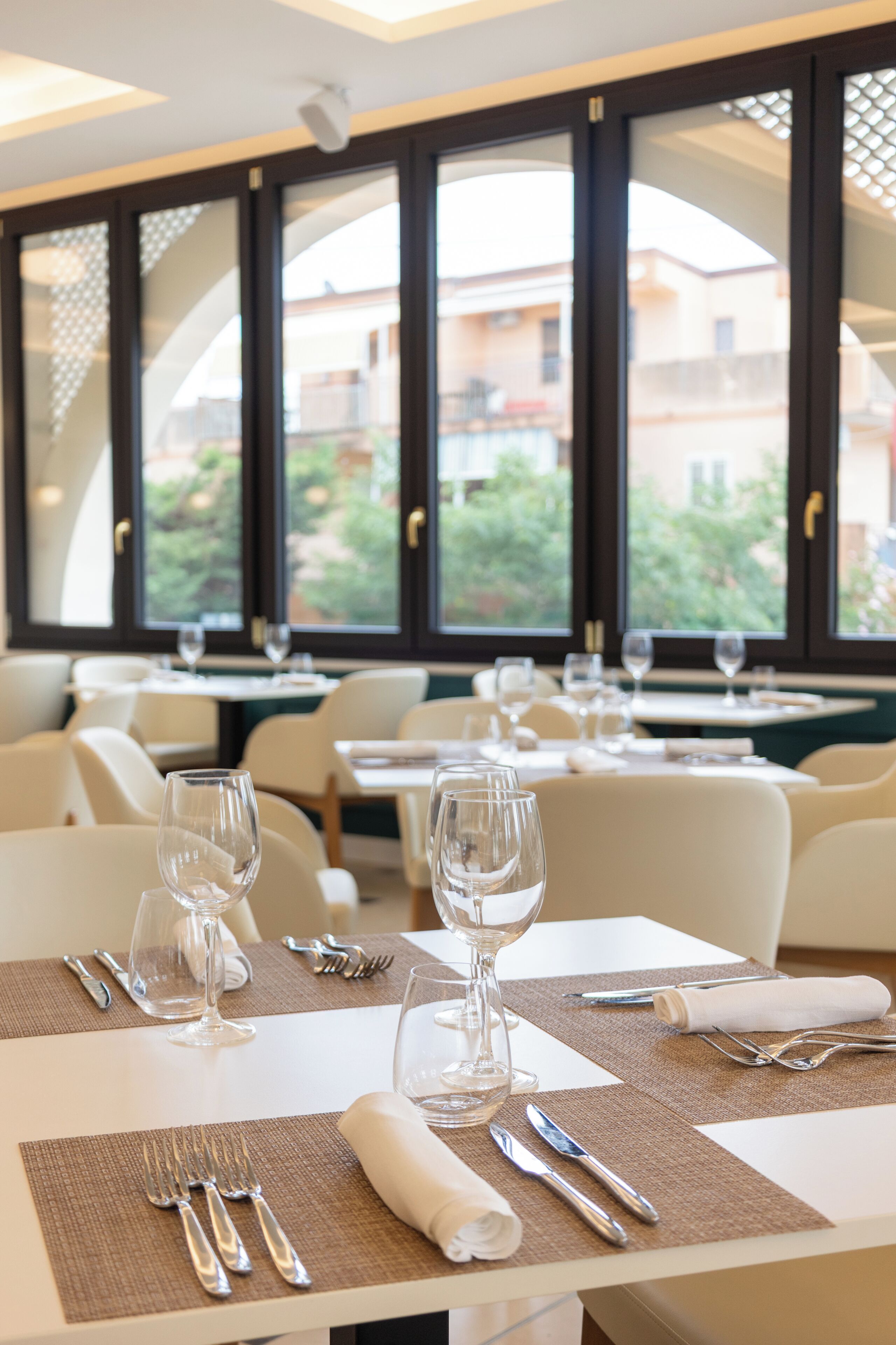 A restaurant interior with tables set for dining, clear glassware, and a view of buildings through large windows.