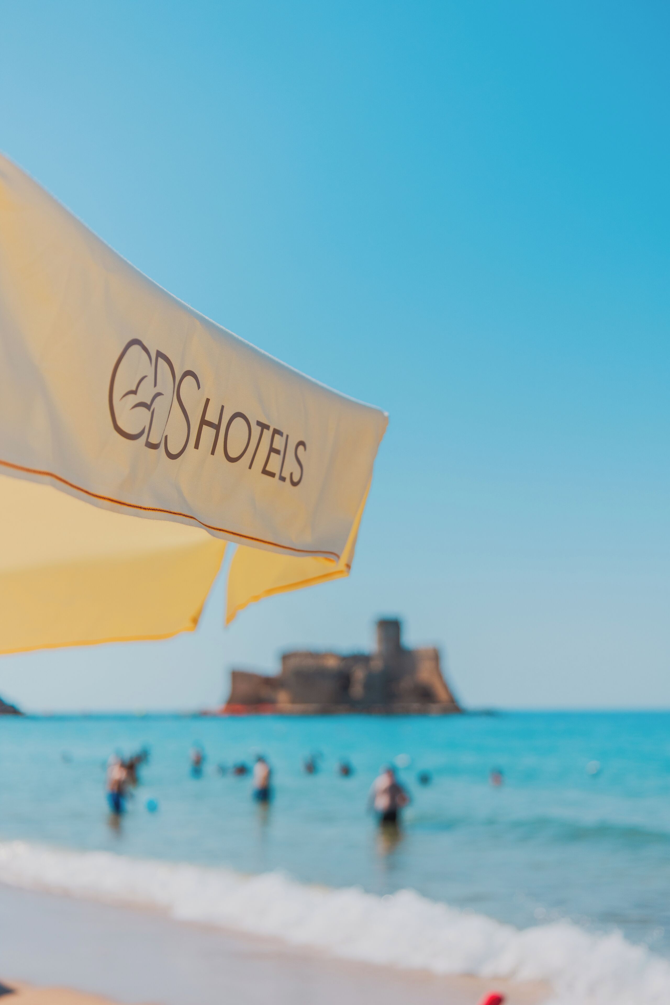 A beach umbrella with the text 'HOTELS' on it, a beach with people in the water, and a distant building on the shore.