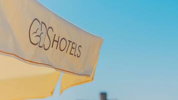 A beach umbrella with the text 'HOTELS' on it, a beach with people in the water, and a distant building on the shore.