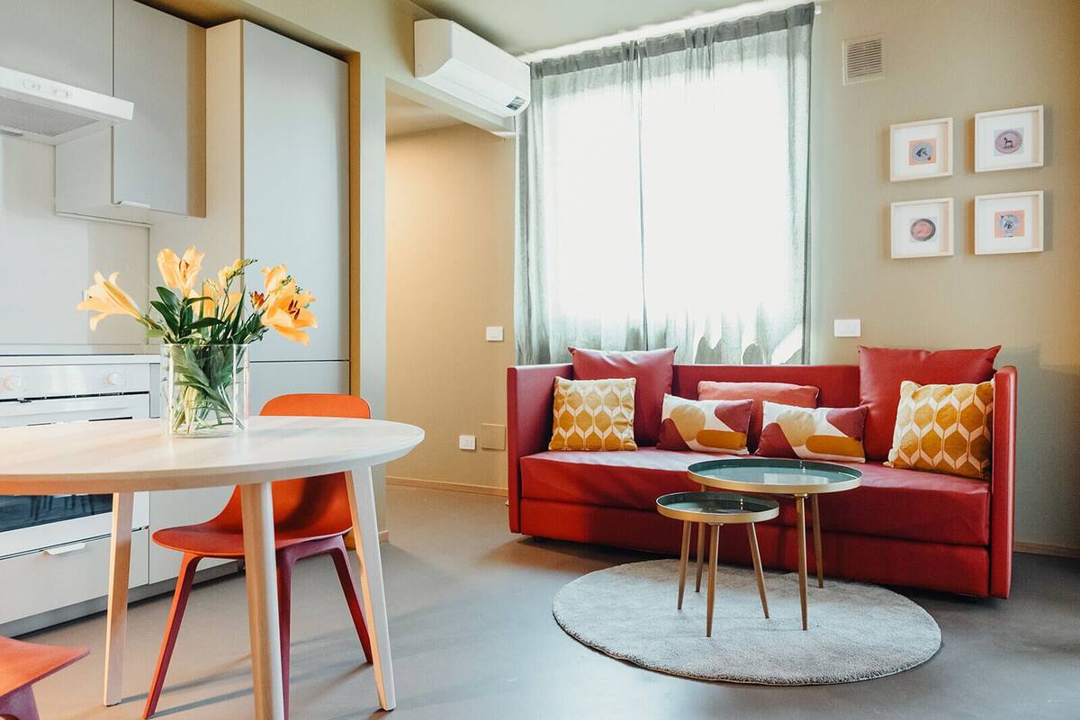 Ein modernes Wohnzimmer mit einem roten Sofa, einem runden Couchtisch und einem Esstisch mit Stühlen. Auf dem Esstisch steht eine Vase mit Blumen.