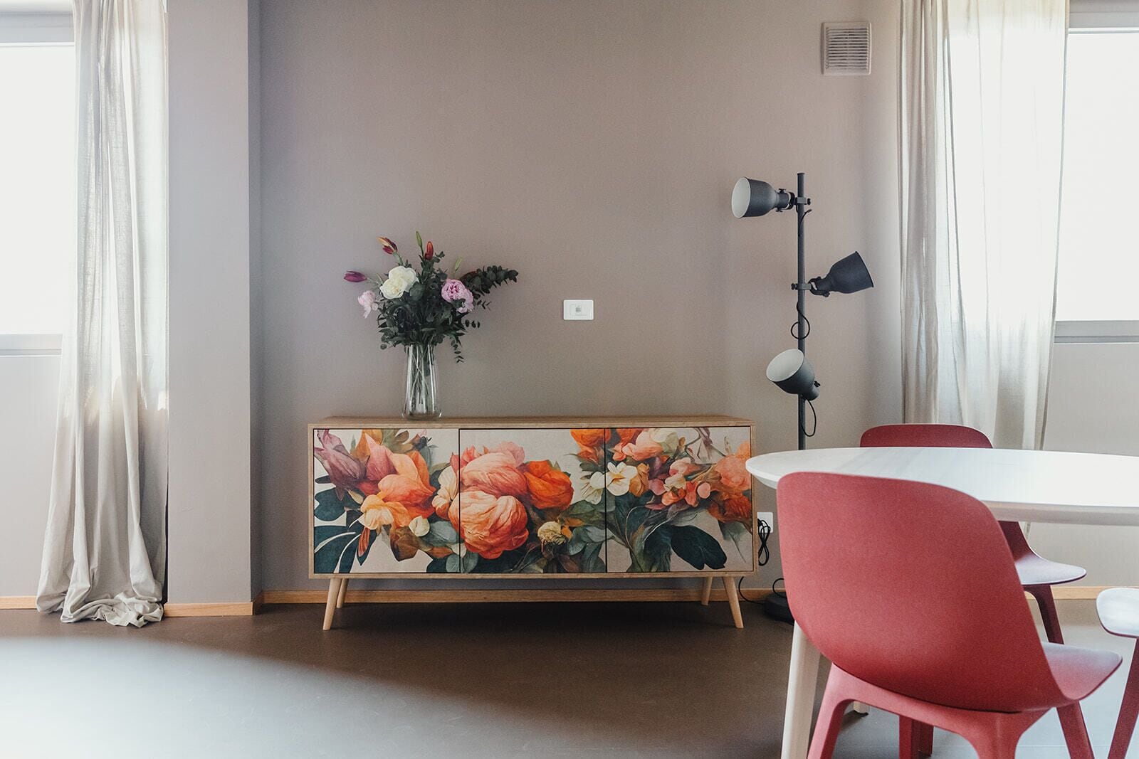 A dining area with a round table, red chairs, a floral cabinet, and a floor lamp.