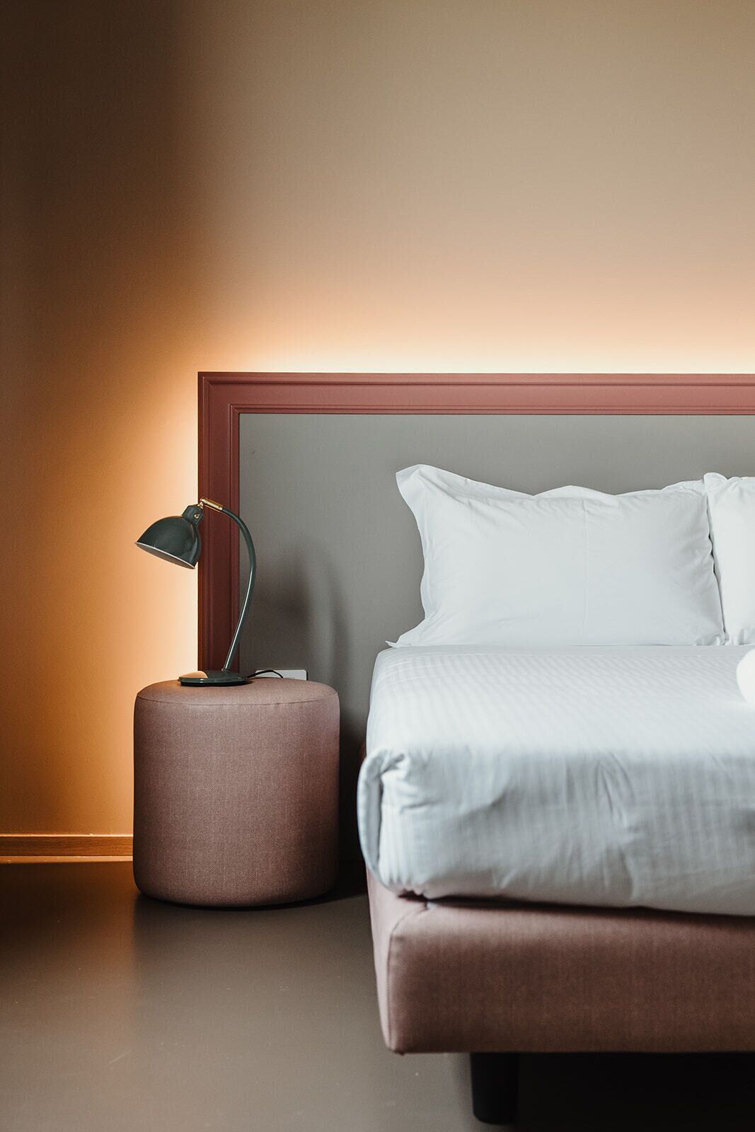 Ein ordentlich bezogenes Bett mit weißen Leinentüchern, ein runder Nachttisch mit einer Lampe und ein Holz-Kopfteil.