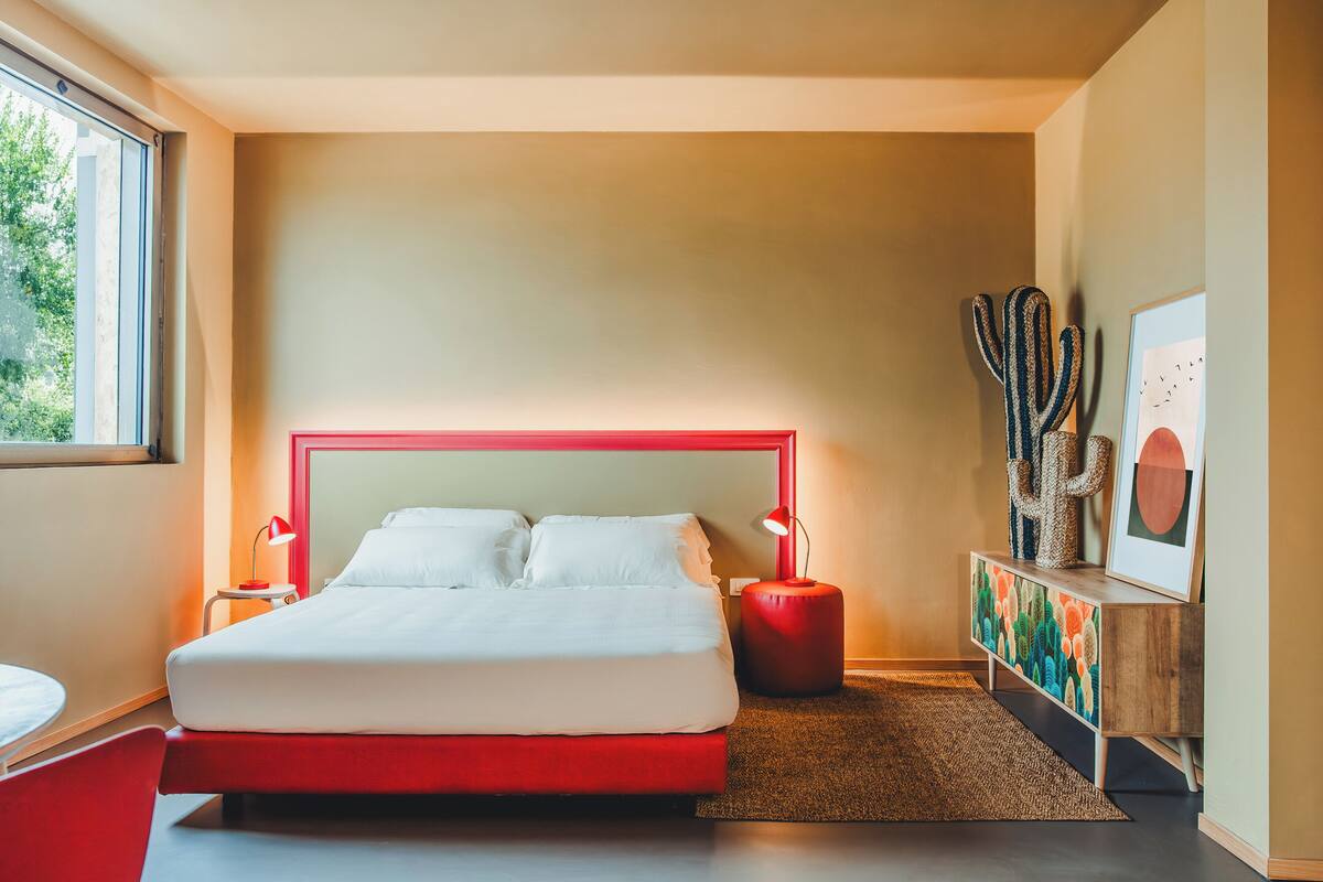 Ein modernes Schlafzimmer mit rotem Bettgestell, weißer Bettwäsche, einer Nachttischlampe, einer Kakteendeko und einem gerahmten Bild auf dem Nachttisch.