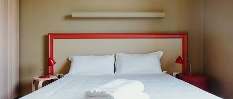 A neatly made bed with white linens and a folded towel in front. There are two bedside tables with red lamps.