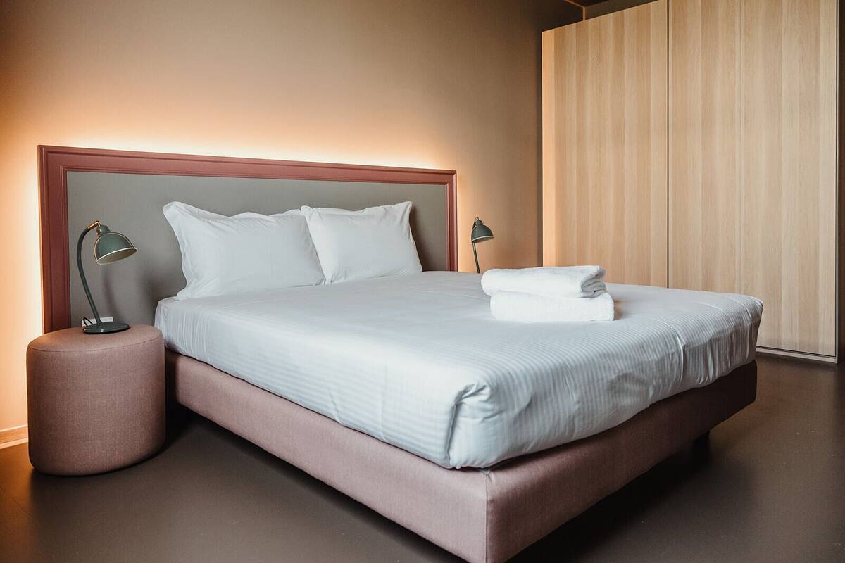Ein ordentlich bezogenes Bett mit weißen Leinen und Kissen, ein runder Nachttisch mit Lampe und eine Wand mit Holzbeschlägen.