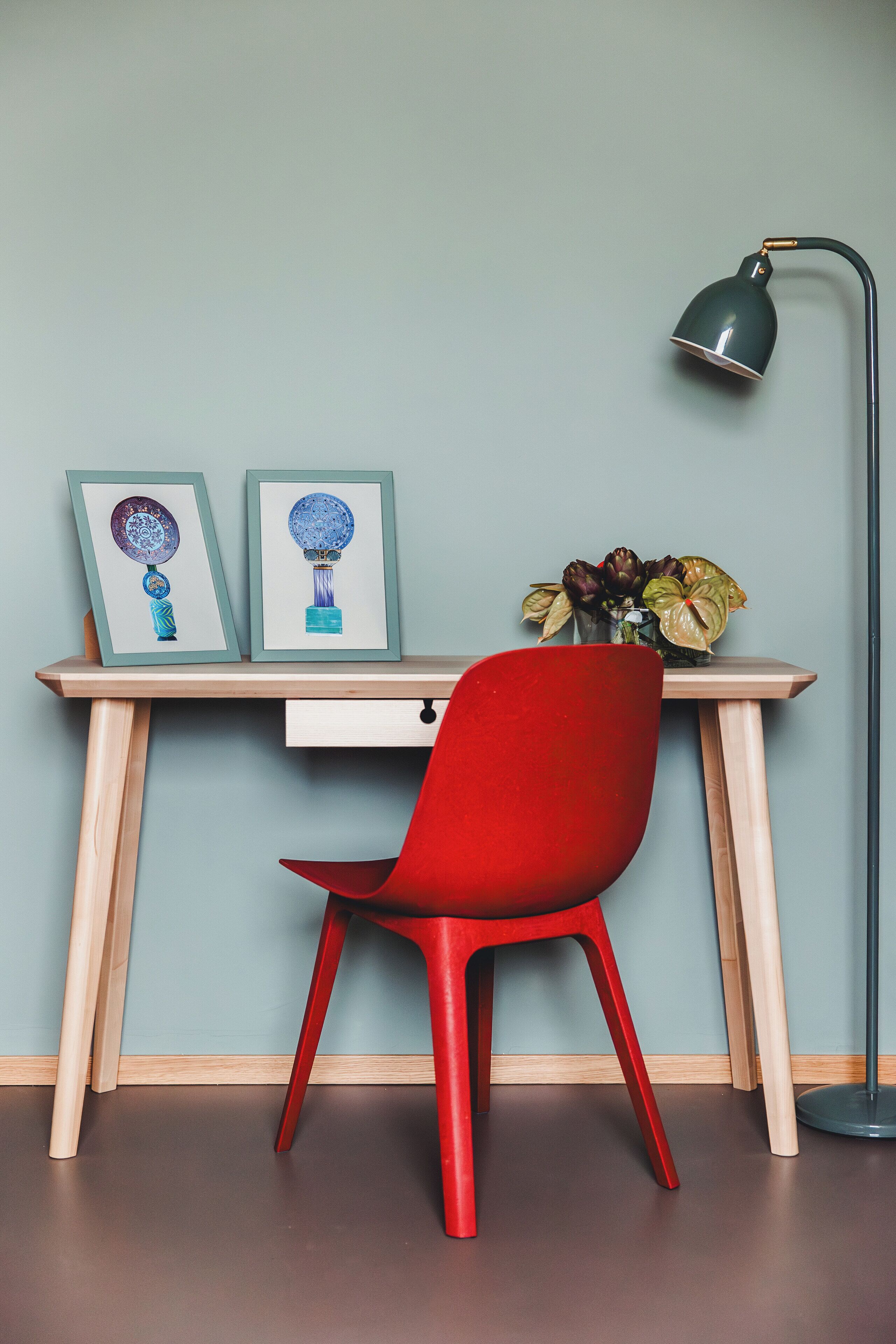 A desk with two framed artworks, a red chair, a floor lamp, and a vase with flowers.