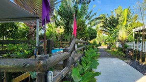 A pathway leading through a tropical garden with a wooden fence, a pool, and various plants.