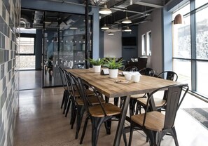 Una sala riunioni moderna con un lungo tavolo di legno, sedie nere, piante in vaso e una parete in vetro con vista sulla città.