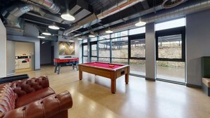 Une table de billard dans une pièce dotée de grandes fenêtres, d’un canapé rouge et d’une table de baby-foot.