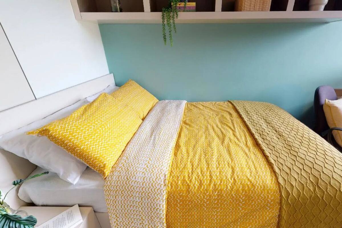 A bed with yellow and white bedding, a desk with a chair, and a hanging plant.