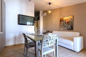 Un salon moderne avec une table à manger en verre, des chaises blanches, un canapé blanc, un téléviseur fixé au mur et un tableau accroché au mur.