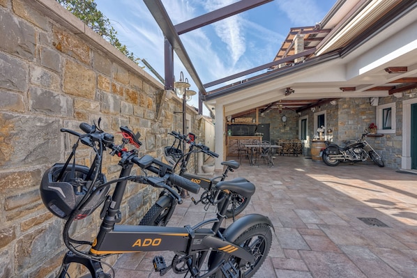 A stone-walled patio with two electric bikes and a motorcycle, a dining area with tables and chairs, and a view of the sky through a glass roof.