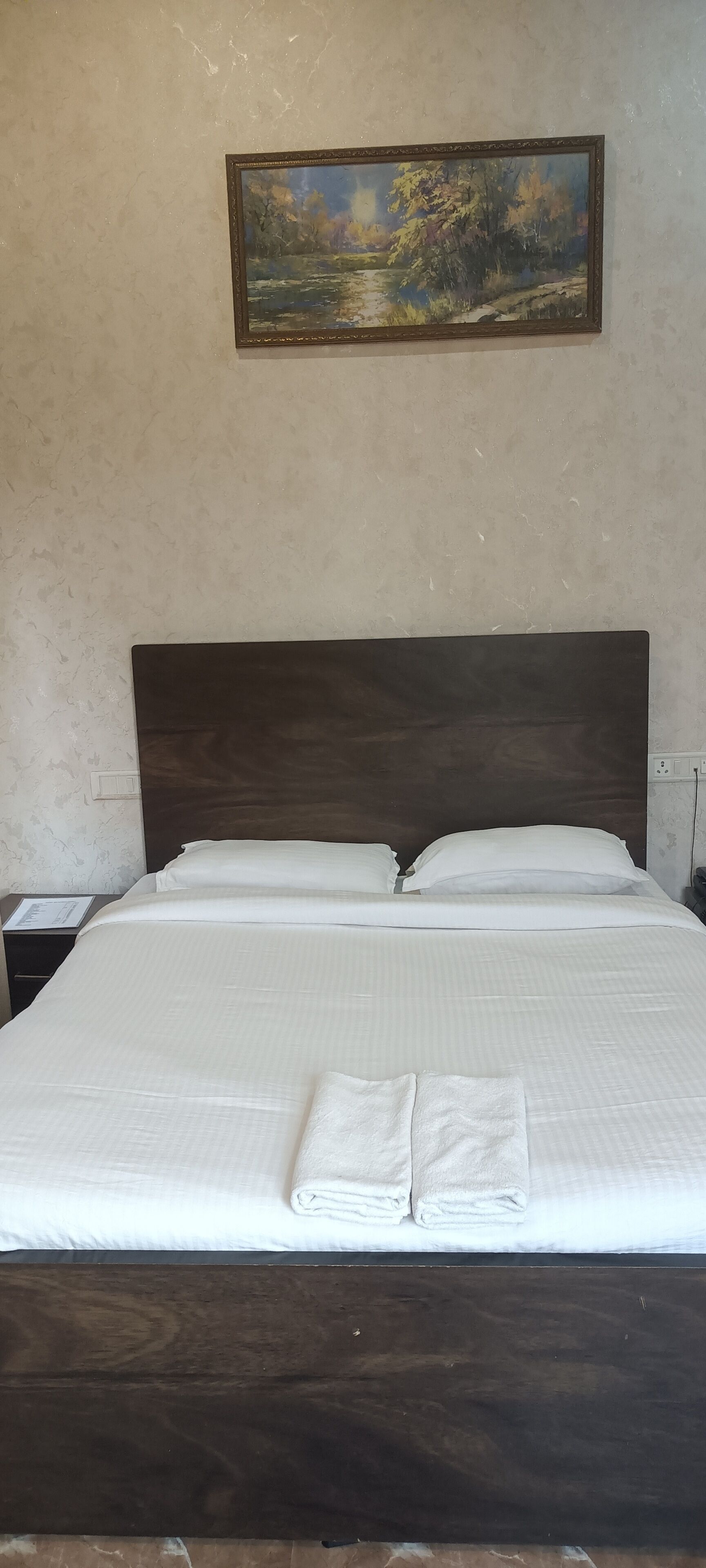 A neatly made bed with white linens and two folded towels, a wooden headboard, and a painting of a landscape on the wall.