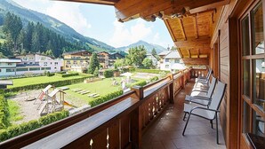 Un balcón de madera con vistas a un paisaje montañoso, un jardín y varios edificios al fondo.