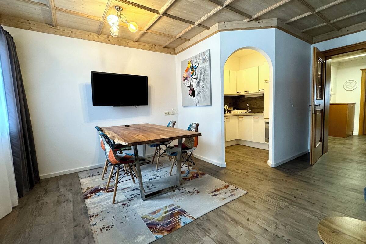 A dining area with a wooden table and chairs, a flat-screen TV mounted on the wall, and a kitchen in the background.