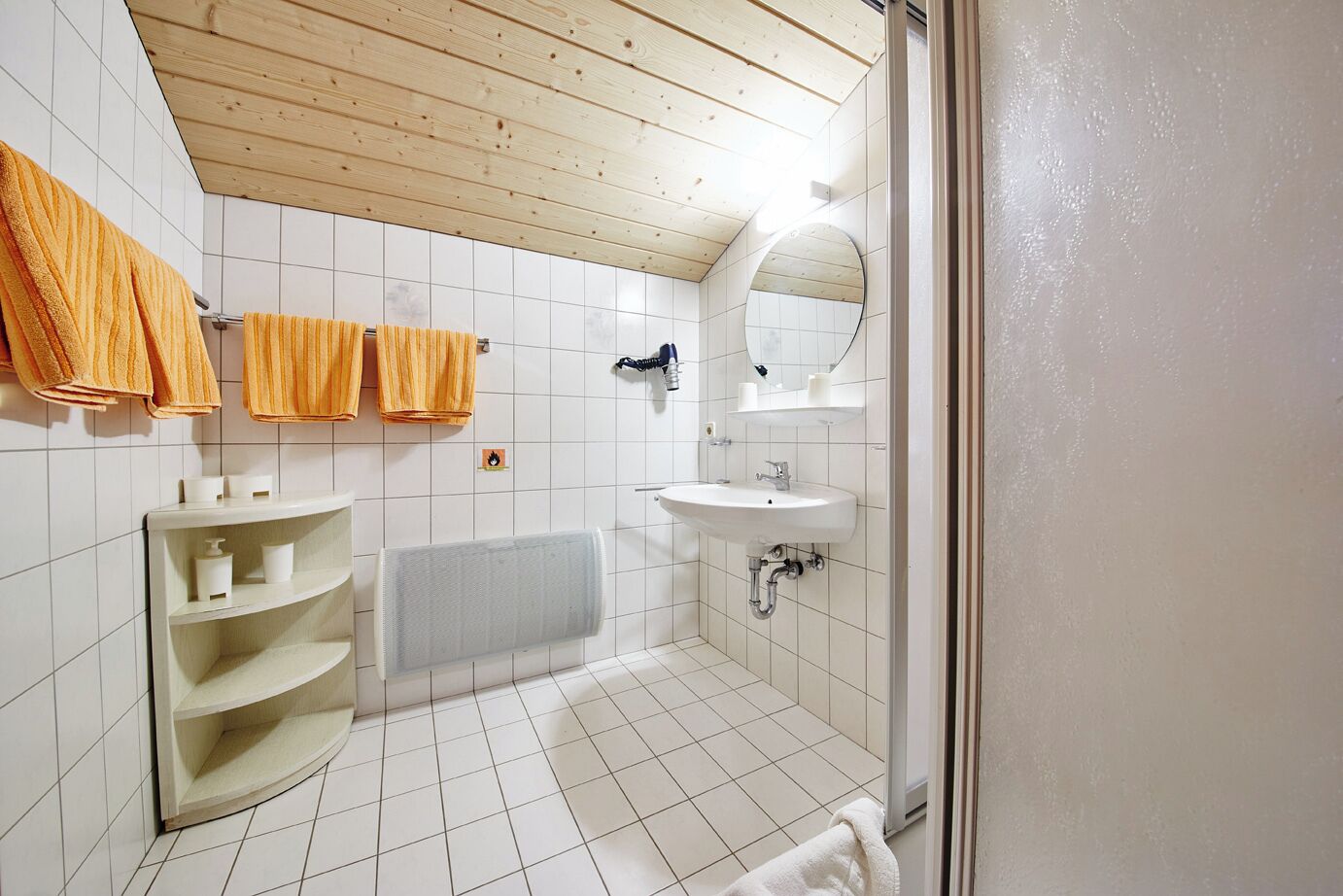 Baño con techo de madera, paredes cubiertas de azulejos blancos, lavamanos, espejo y dos toalleros con toallas.