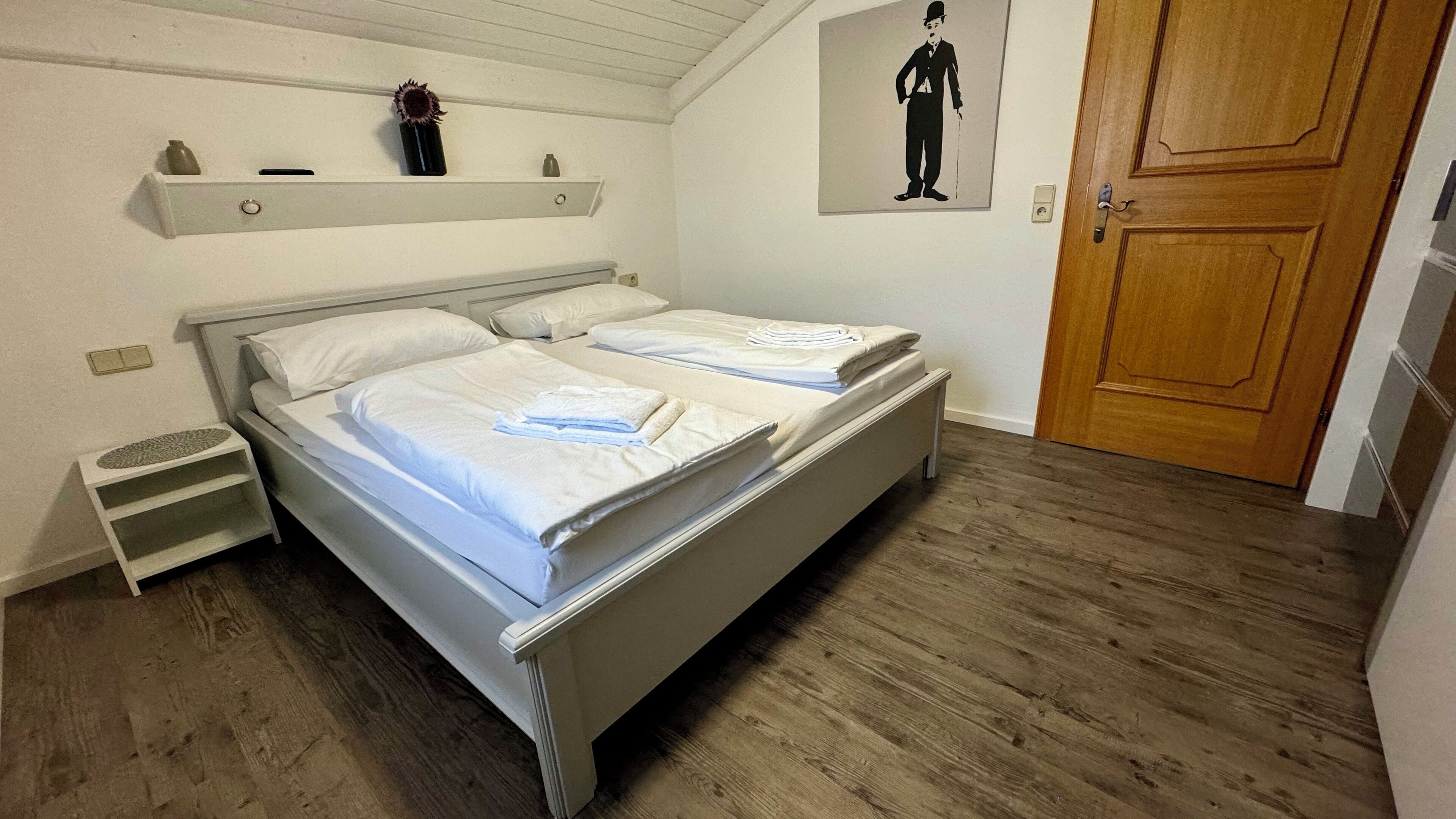 Ein Doppelbett mit weißer Bettwäsche, Holzboden, Holztür und ein an der Wand befestigtes Regal mit einer Vase und einem Bild.