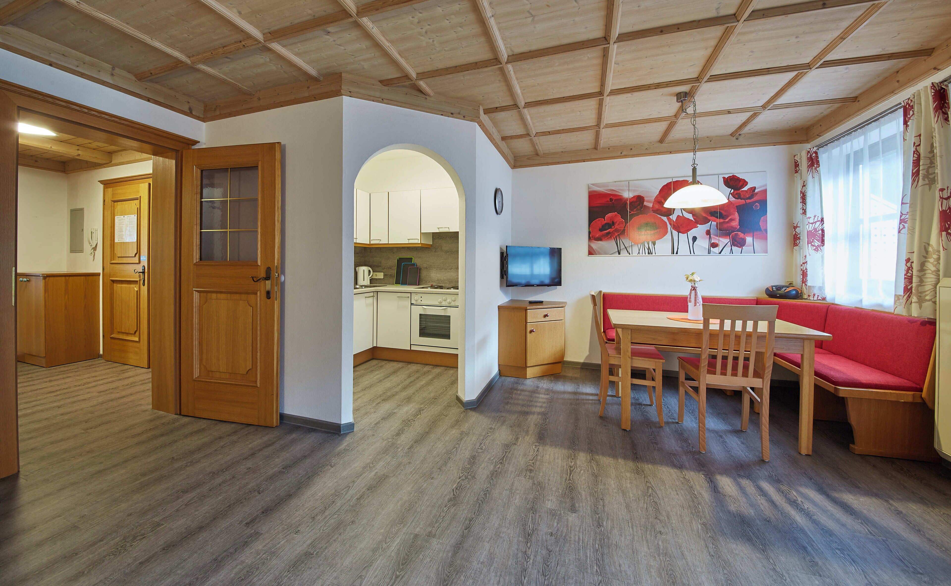 A cozy living room with a wooden ceiling, a dining area with a red sofa and wooden table, and a kitchen with white cabinets and a TV.