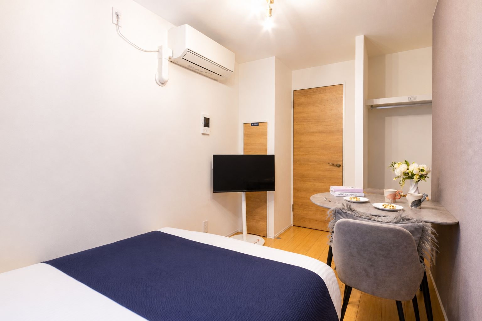 Habitación de hotel compacta con cama, televisor de pantalla plana, una mesita con un jarrón de flores y una puerta de madera.